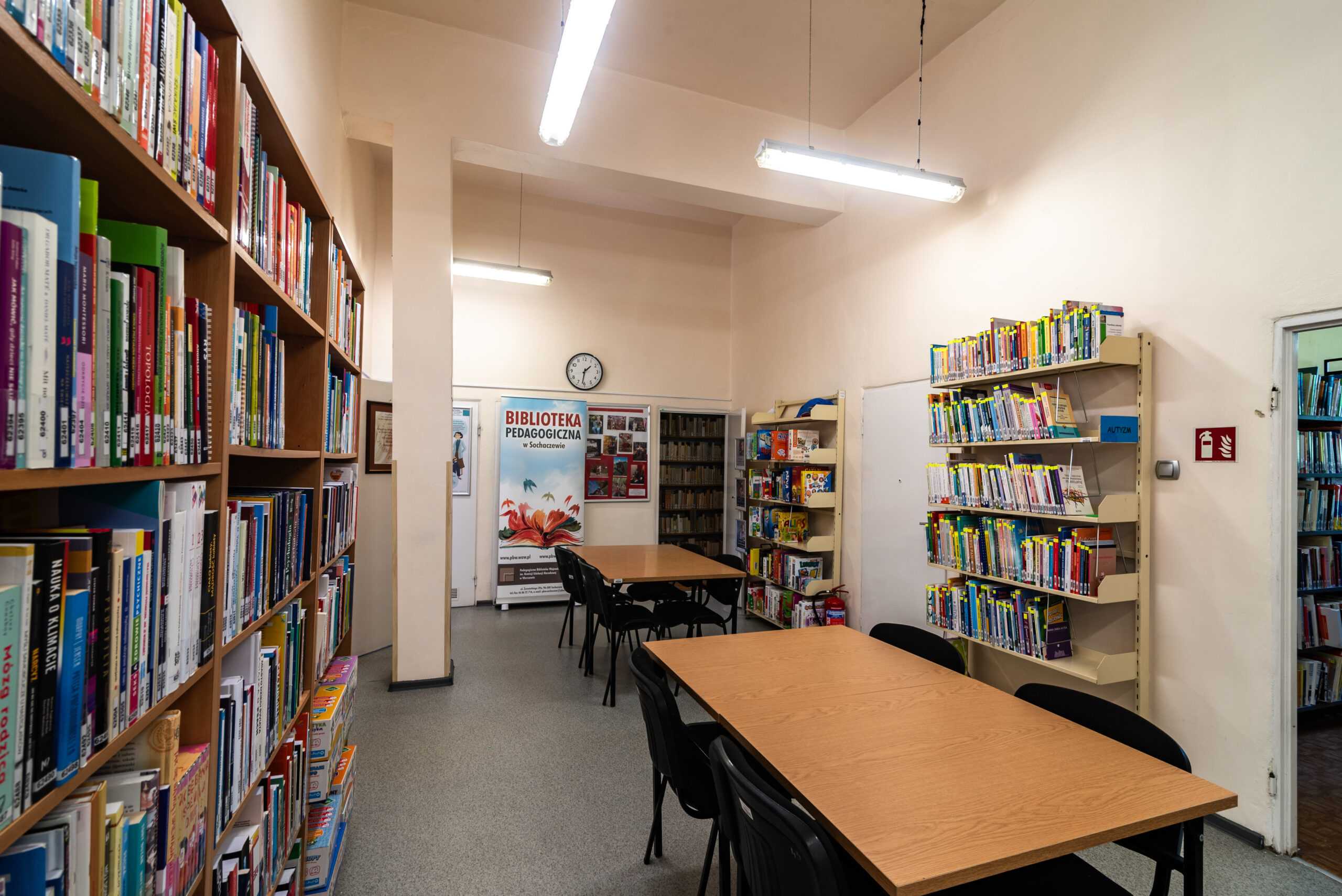 Czytelnia w bibliotece w Sochaczewie. W centralnej części znajdują się dwa duże, drewniane stoły z czarnymi krzesłami. Ściany są jasne, a pod wysokim sufitem wiszą podłużne lampy jarzeniowe. Po lewej stronie i w głębi stoją wysokie regały z książkami. Na wprost widoczny jest banner informacyjny biblioteki oraz tablica z ogłoszeniami. Wnętrze ma klasyczny, naukowy charakter